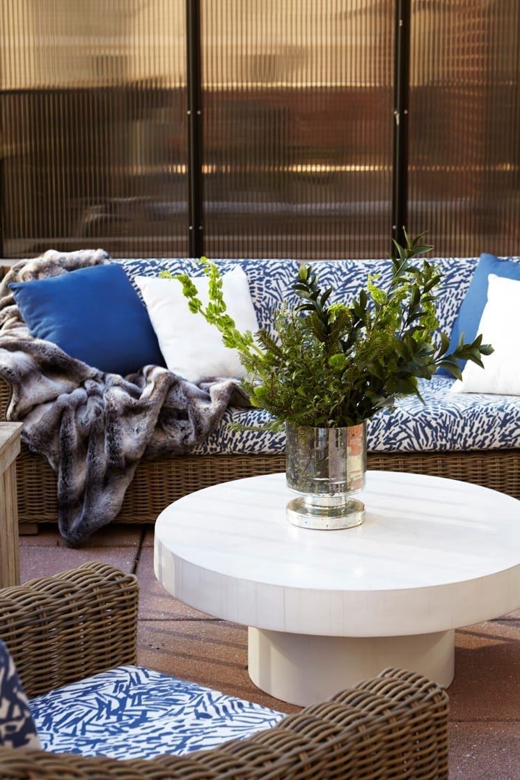 A couch and chair on the terrace of the Gardens Suite Hotel. A large round coffee table is placed at the middle, holding a flower vase. A fuzzy blanket has been thrown over one side of the couch.