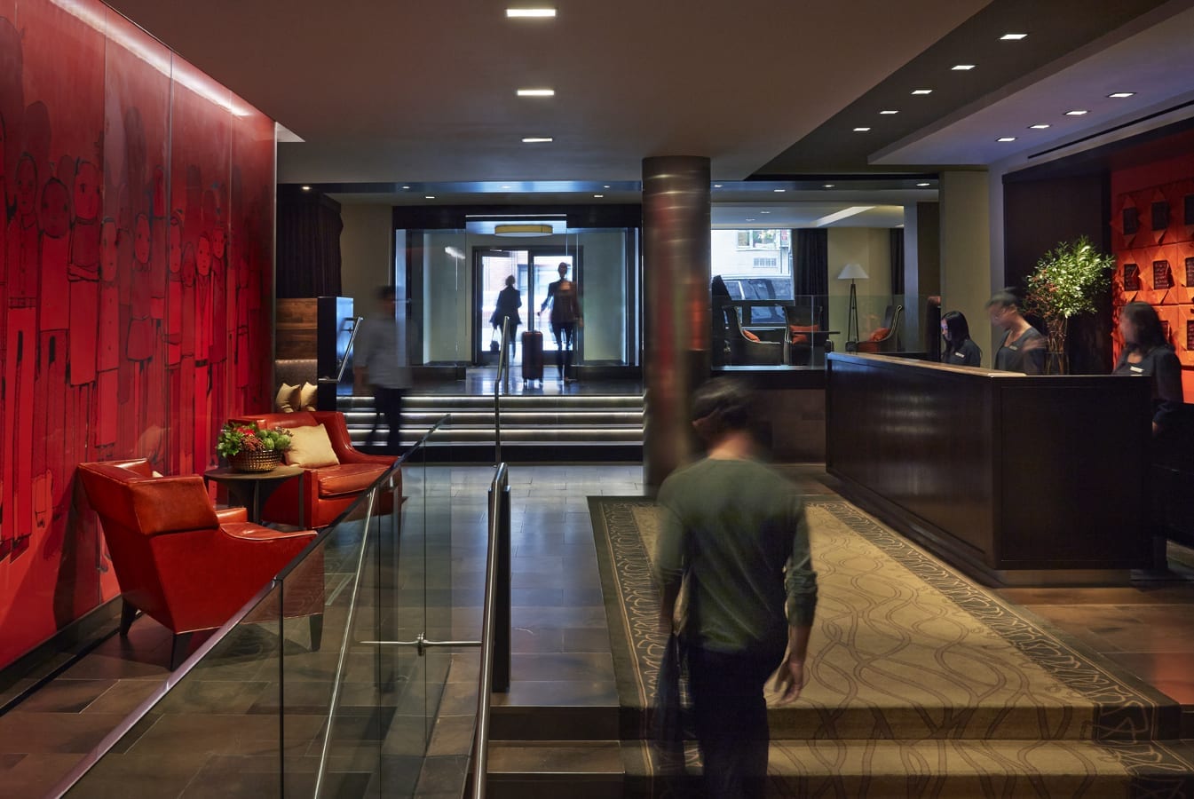 A man walks up the stairs leading to Shelburne Hotel and Suites' front desk. Across at the left are two red chairs against a red wall dressed with texture.