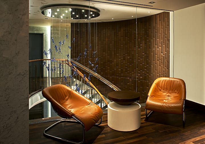 View of a lounge area of the Fifty Hotel and Suites. A curved stretched staircase can be seen behind. Two chairs are placed by the railing, with a round table placed between.